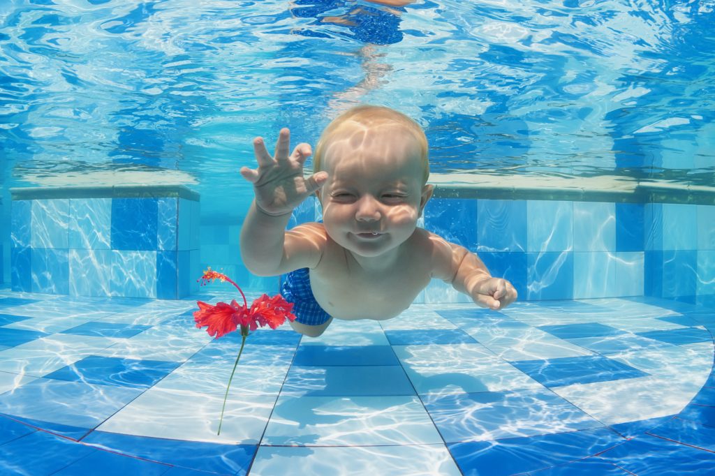 Smiling baby boy diving underwater with fun for red flower in blue pool Active lifestyle, child swimming lesson with parents,